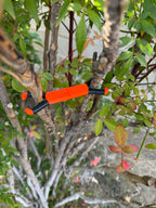 Shine Spine Branch Spreader Solar Ember Orange 2inch gardening tool attached to a tree branch with green leaves in the background.