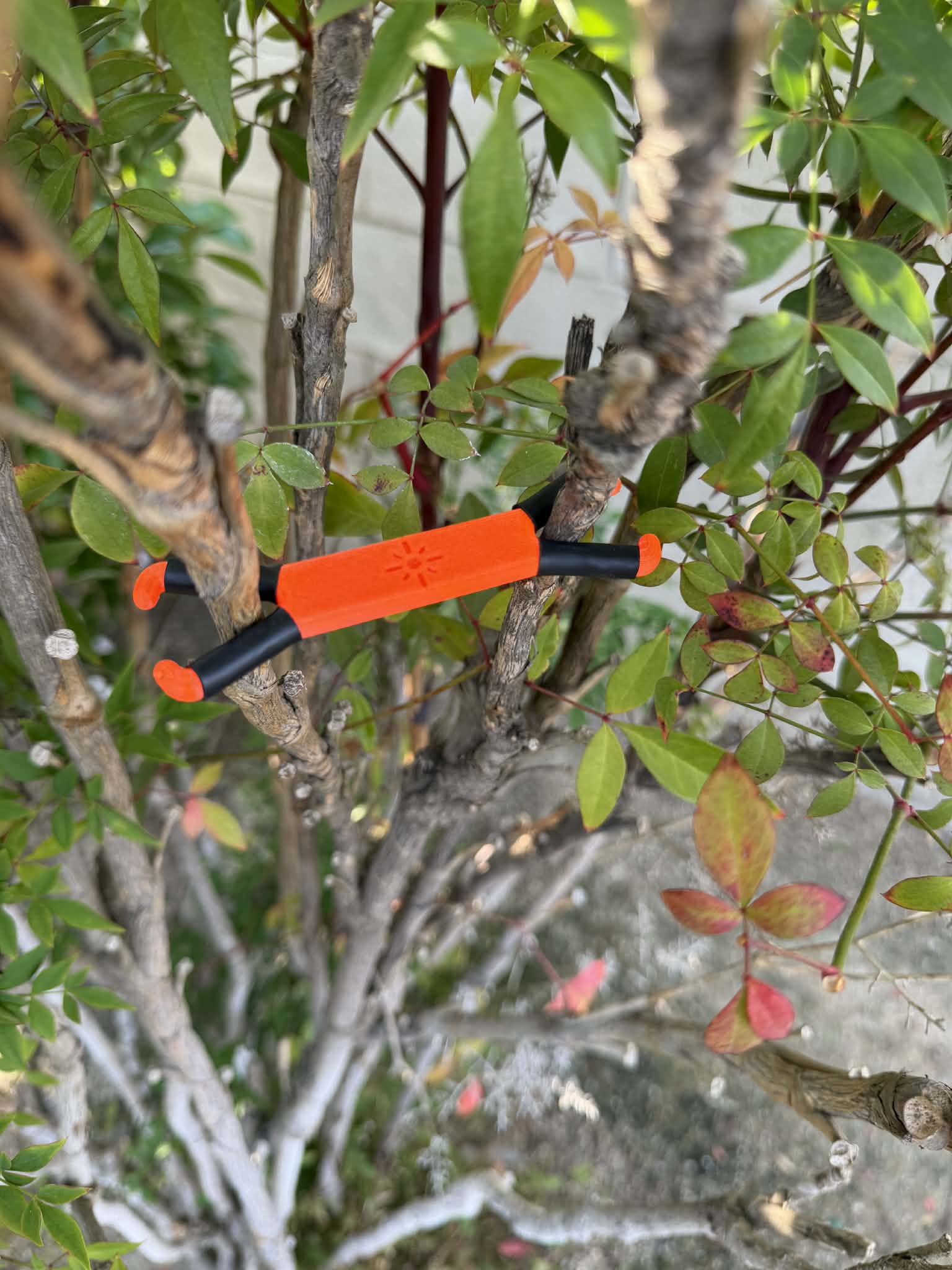 Shine Spine Branch Spreader Solar Ember Orange 2inch gardening tool attached to a tree branch with green leaves in the background.